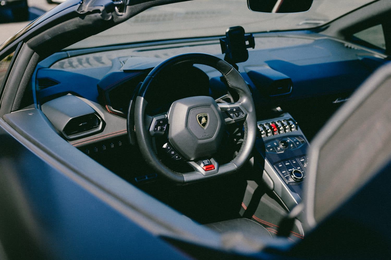 Huracán EVO cockpit through the driver window: flat-bottom steering wheel, dark leather with red contrast stitching, switch-panel console