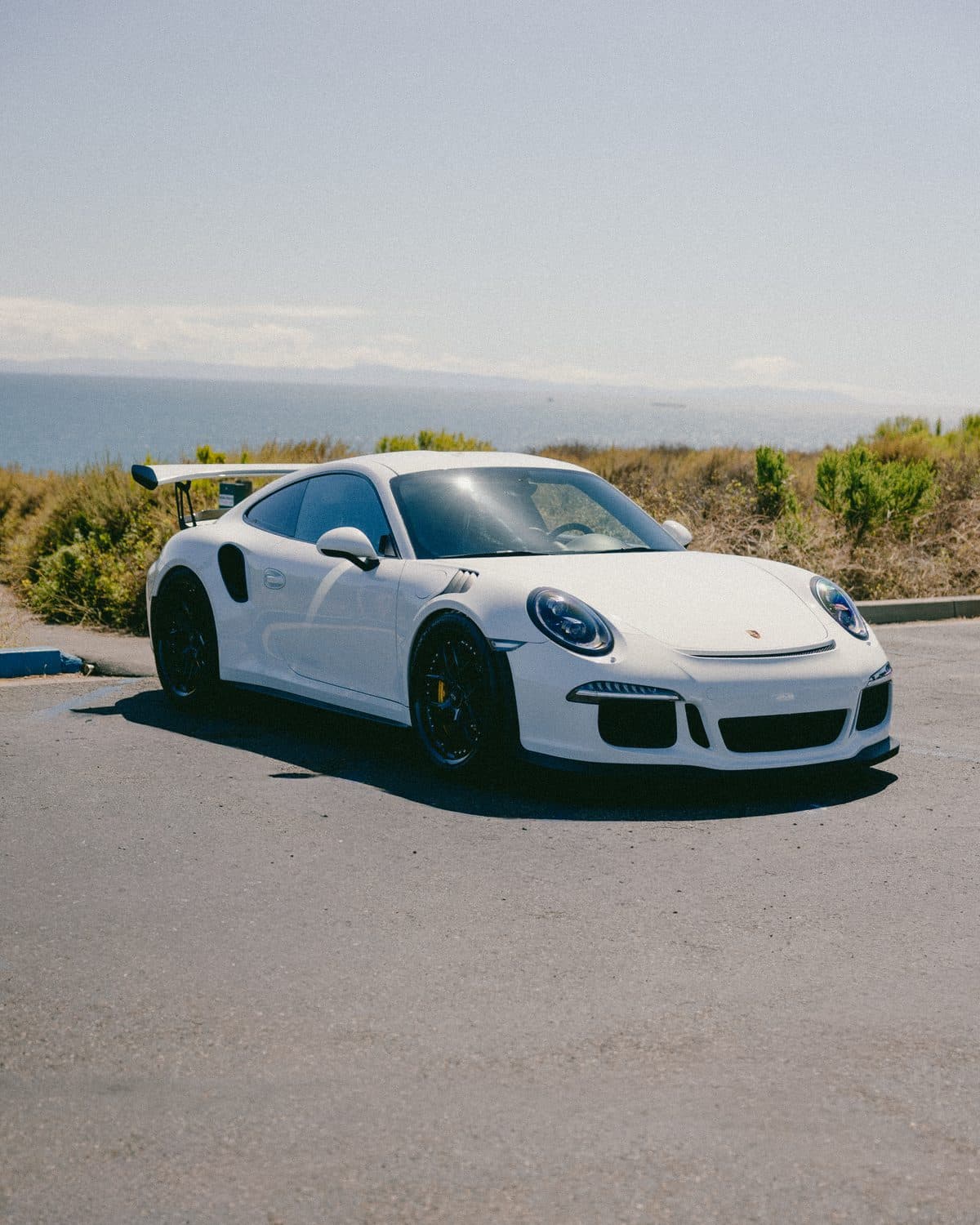 White Porsche 911 GT3 RS from the passenger-side front three-quarter, side intakes and rear wing visible, hazy coastline behind