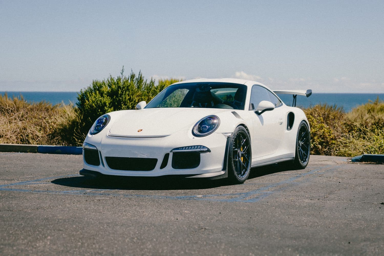 White Porsche 911 GT3 RS from the driver-side front three-quarter, black center-lock wheels and fixed rear wing, coast and ocean behind