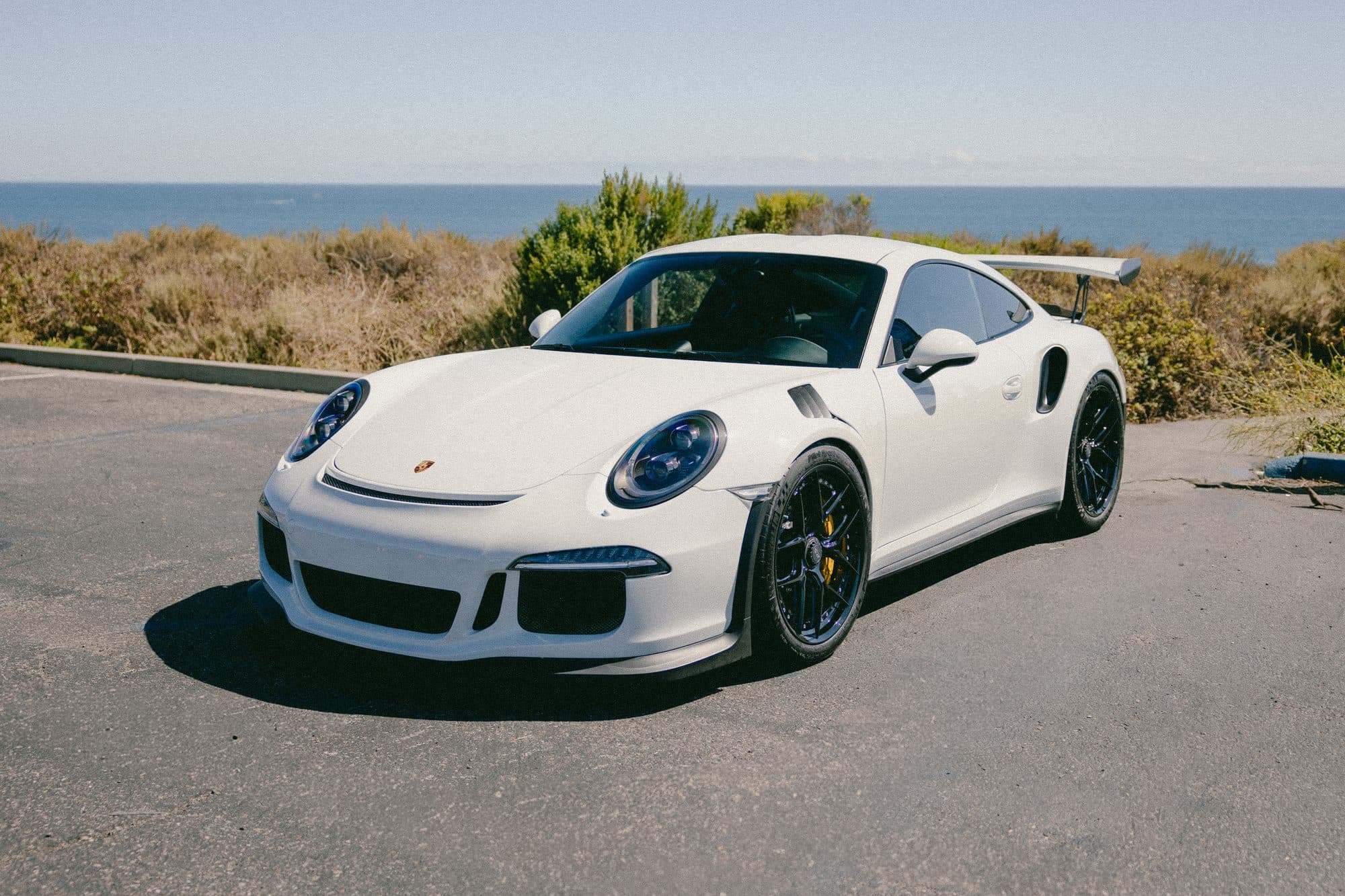 White Porsche 911 GT3 RS at a front three-quarter angle in an oceanfront lot, black wheels and rear wing visible, Pacific horizon behind